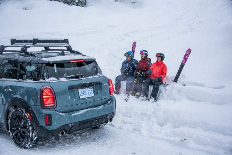 Image of three “Silver Sliders”, who are sitting in the snow with their ski equipment next to a MINI Countryman.  