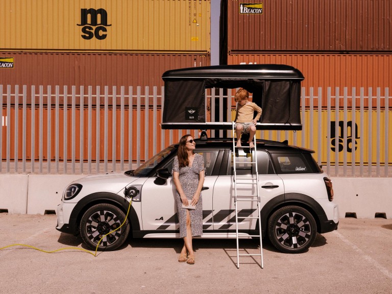 MINI Countryman припаркован перед грузовыми контейнерами и заряжается на зарядной станции. Пепе сидит в палатке на крыше, его мама стоит перед автомобилем.